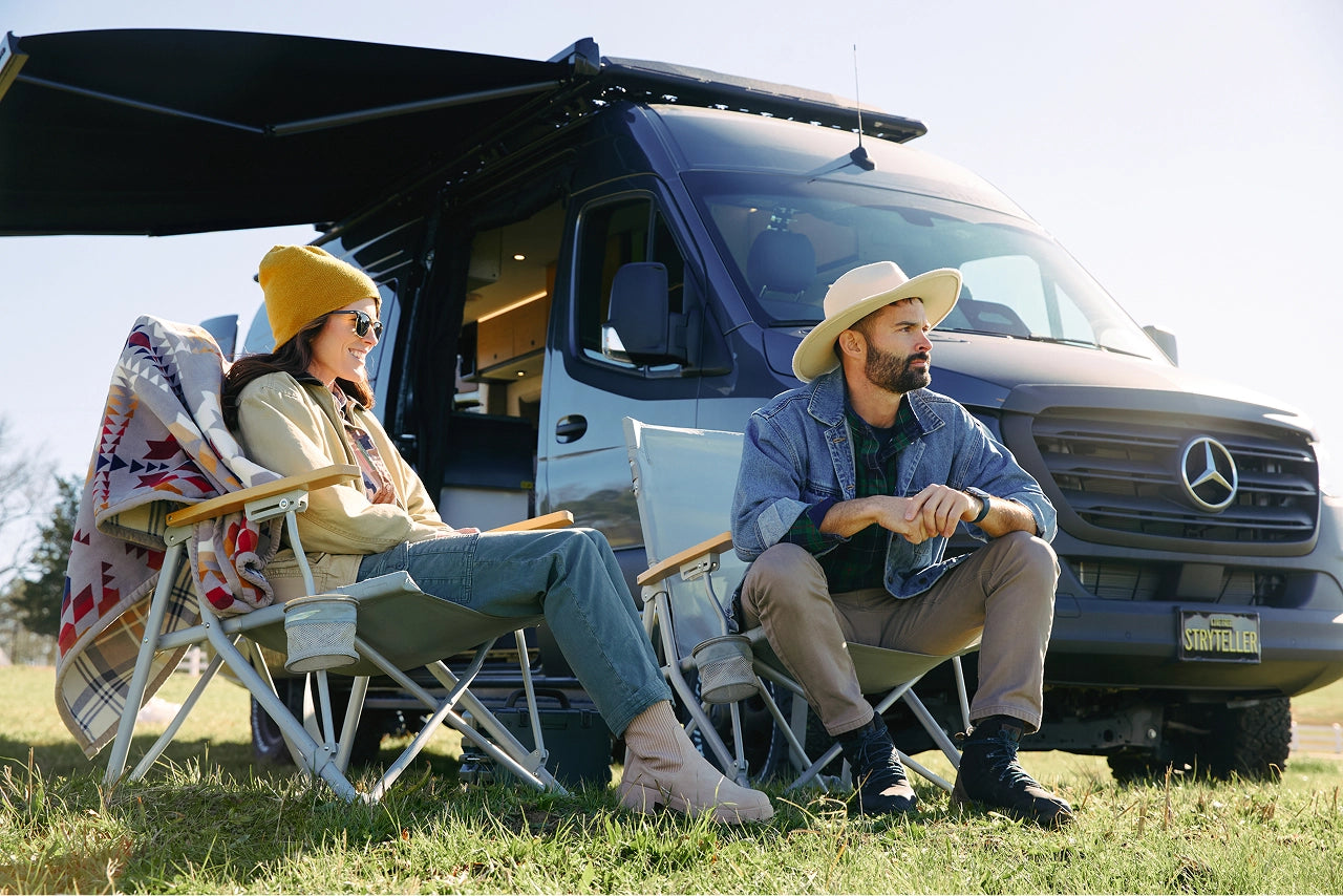 Two people sitting in front of a Mercedes-Benz camper van in a grassy area. Storyteller Overland, Camper Van, Campervan, Adventure Van, Class B RV, Small Motorhome, Overland, Overlanding, Offroad Vehicle, Vanlife, Van life Awning