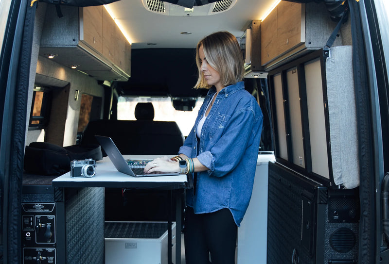 Woman working on laptop inside custom camper van conversion, featuring modern cabinetry, ideal for vanlife and adventure travel.