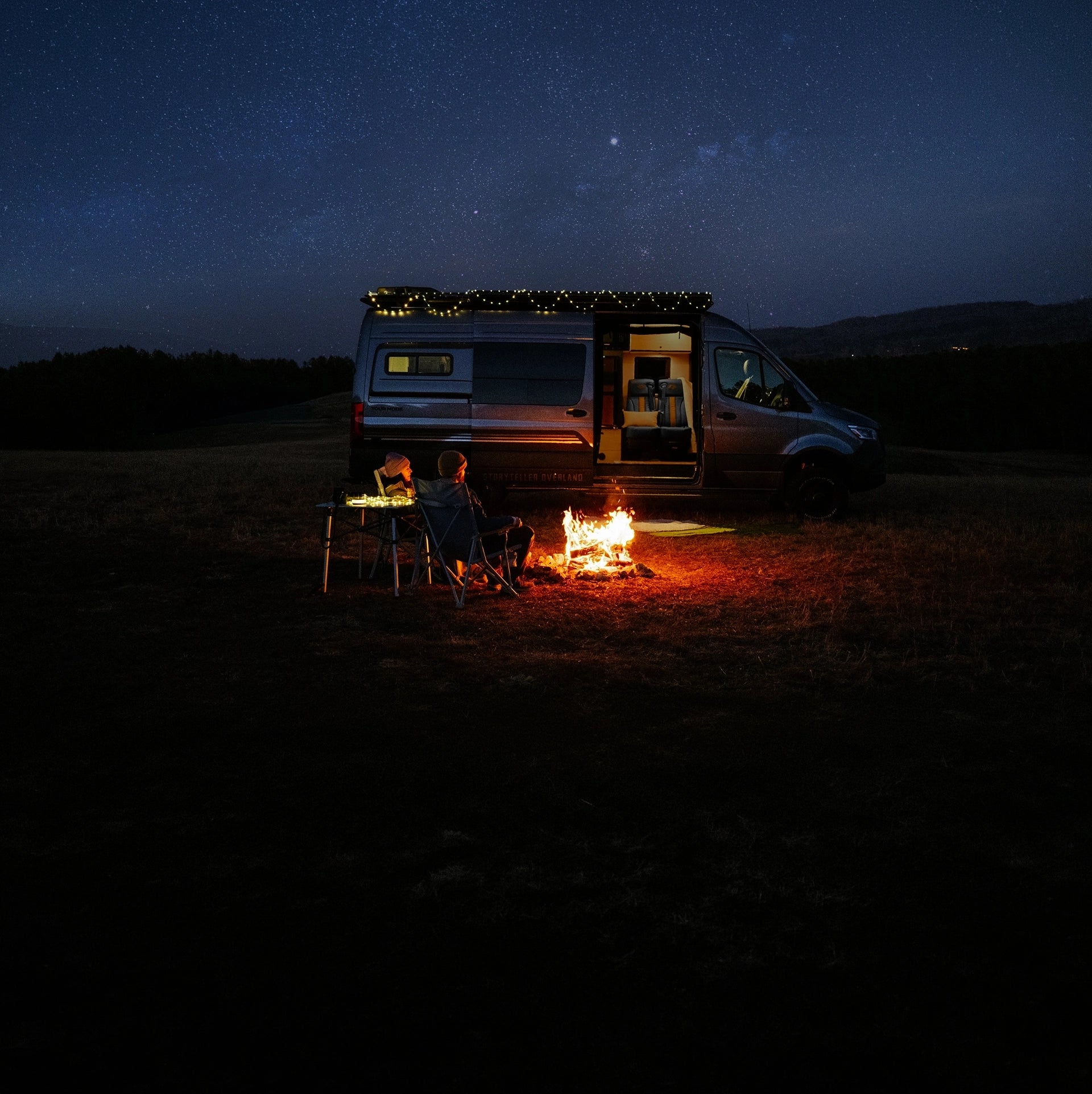 Storyteller Overland, Camper Van, Campervan, Adventure Van, Class B RV, Small Motorhome, Overland, Overlanding, Offroad Vehicle, Vanlife, Van life, TOUR MODE. Two people sitting by a campfire in front of a camper van under a starry night sky.