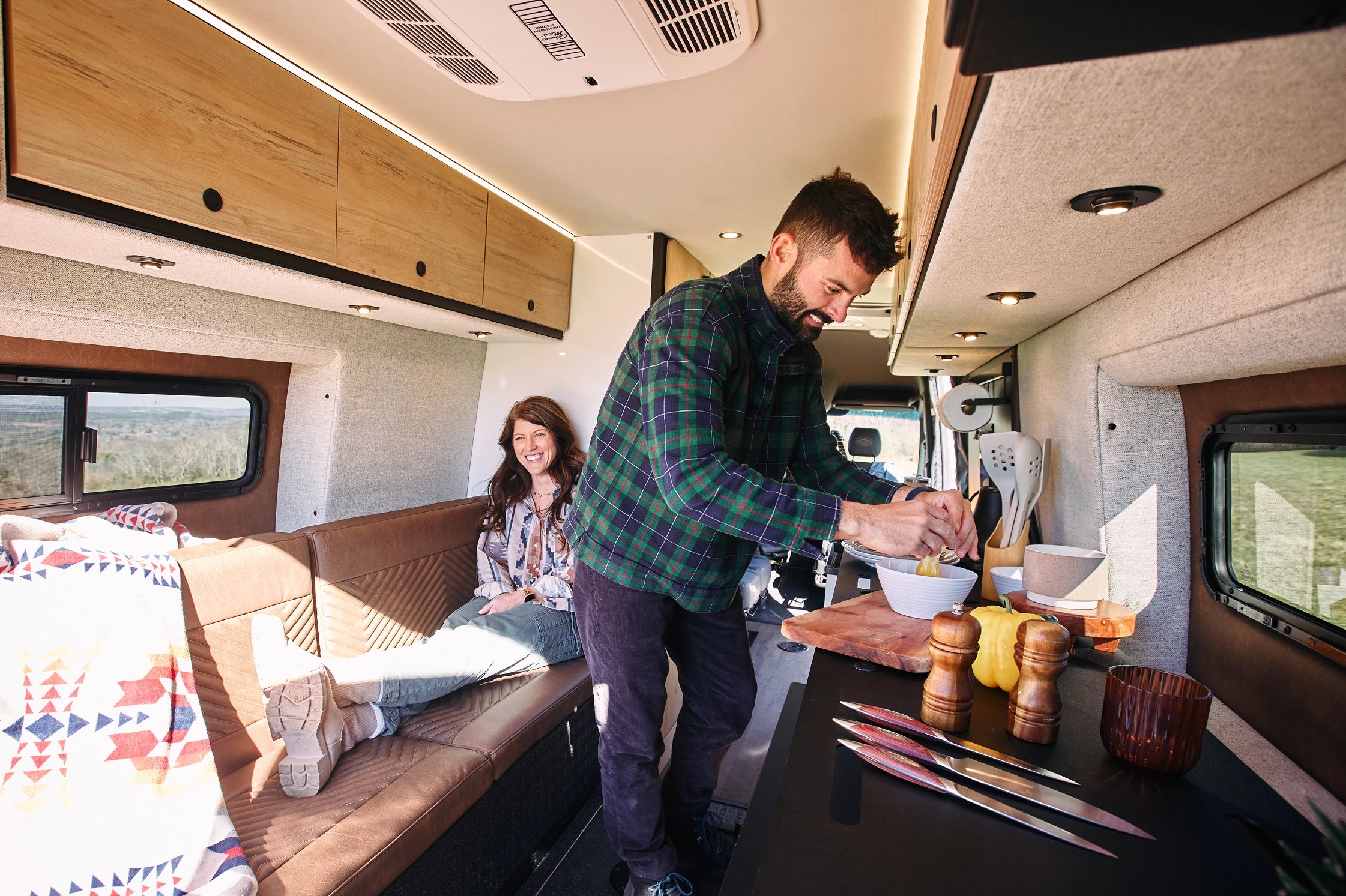 Man and woman inside a camper van preparing food together. Camping Storyteller Overland, Camper Van, TOUR MODE Mercedes Sprinter