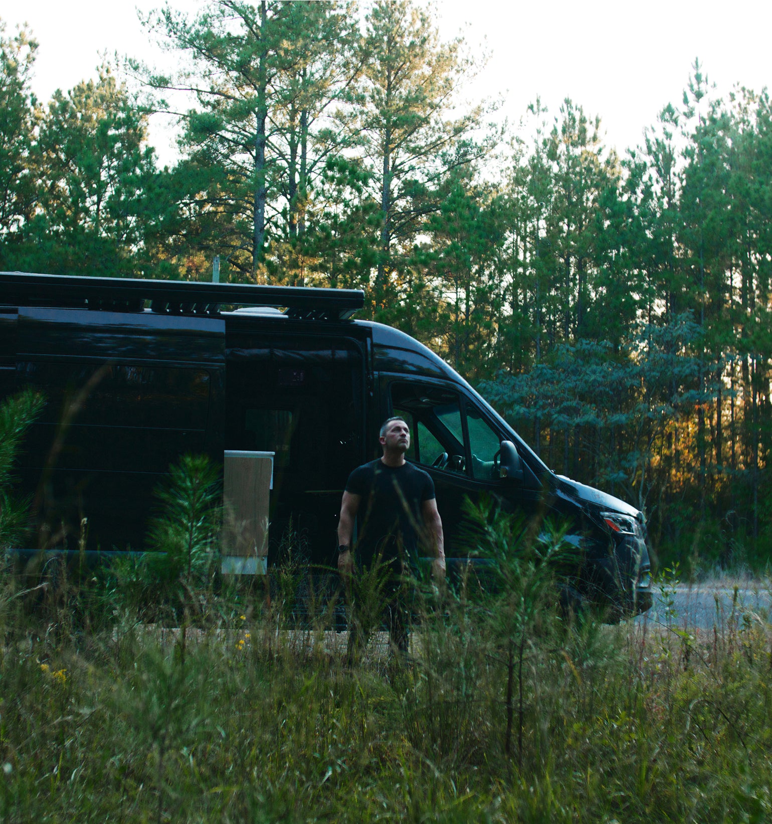 Man stands by black Sprinter 4x4 camper van in forest, overland adventure rig, vanlife setting
