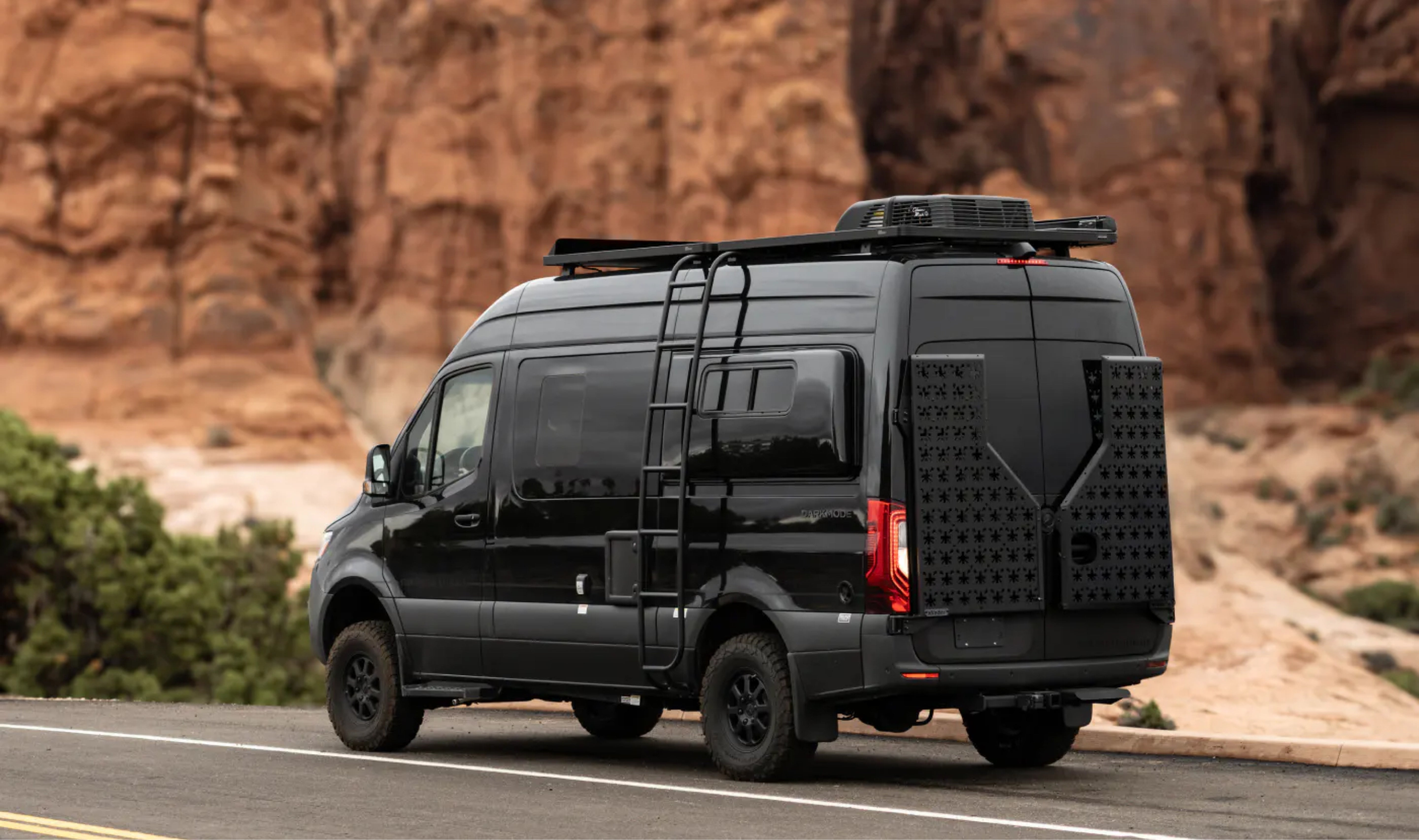 Black Sprinter 4x4 adventure van with ladder and roof rack parked in desert landscape, perfect for vanlife, camper van, overlanding, and road trip travel.