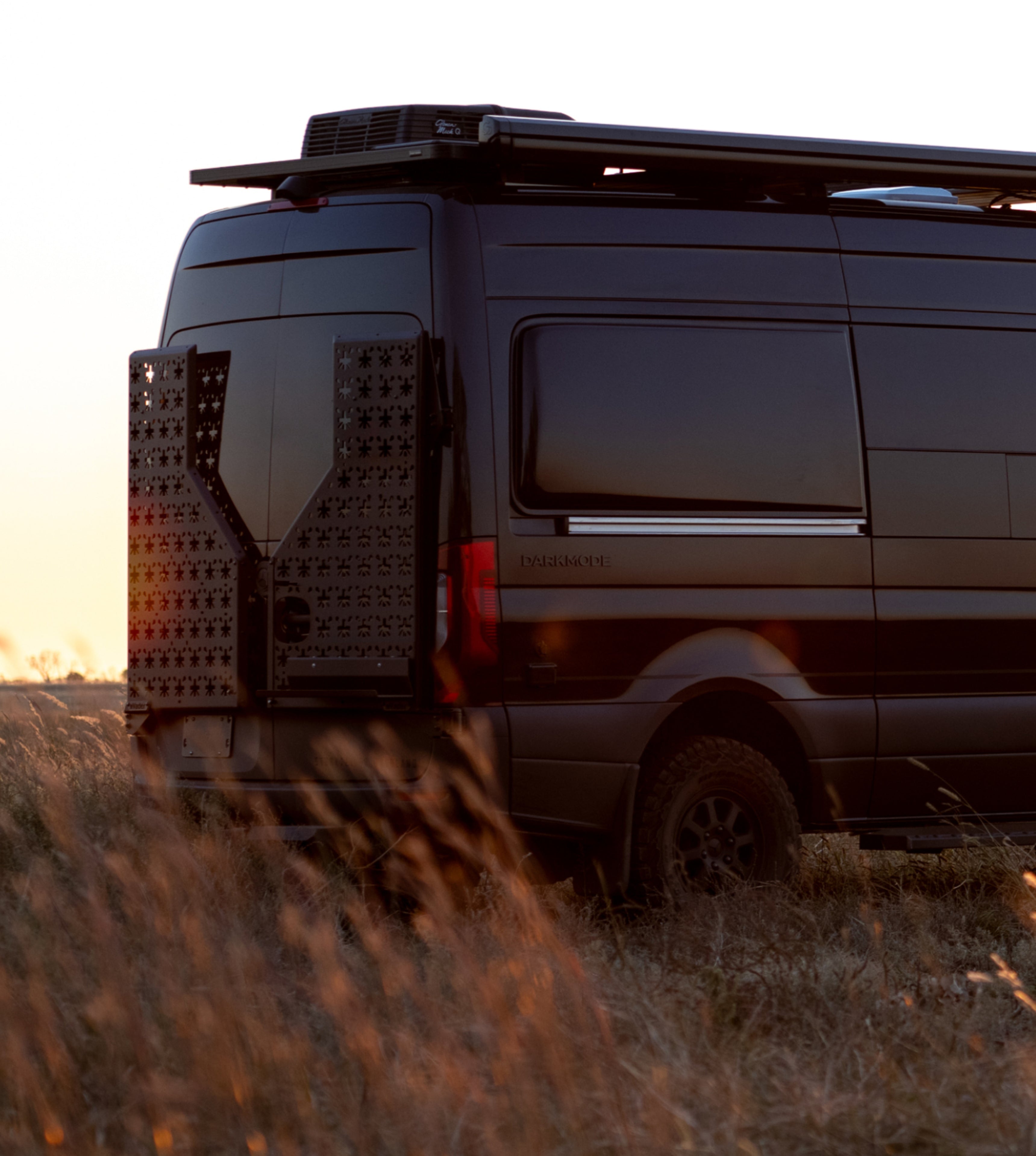 Dark stealth camper van with offroad tires and rear gear on grass, adventure vanlife setting