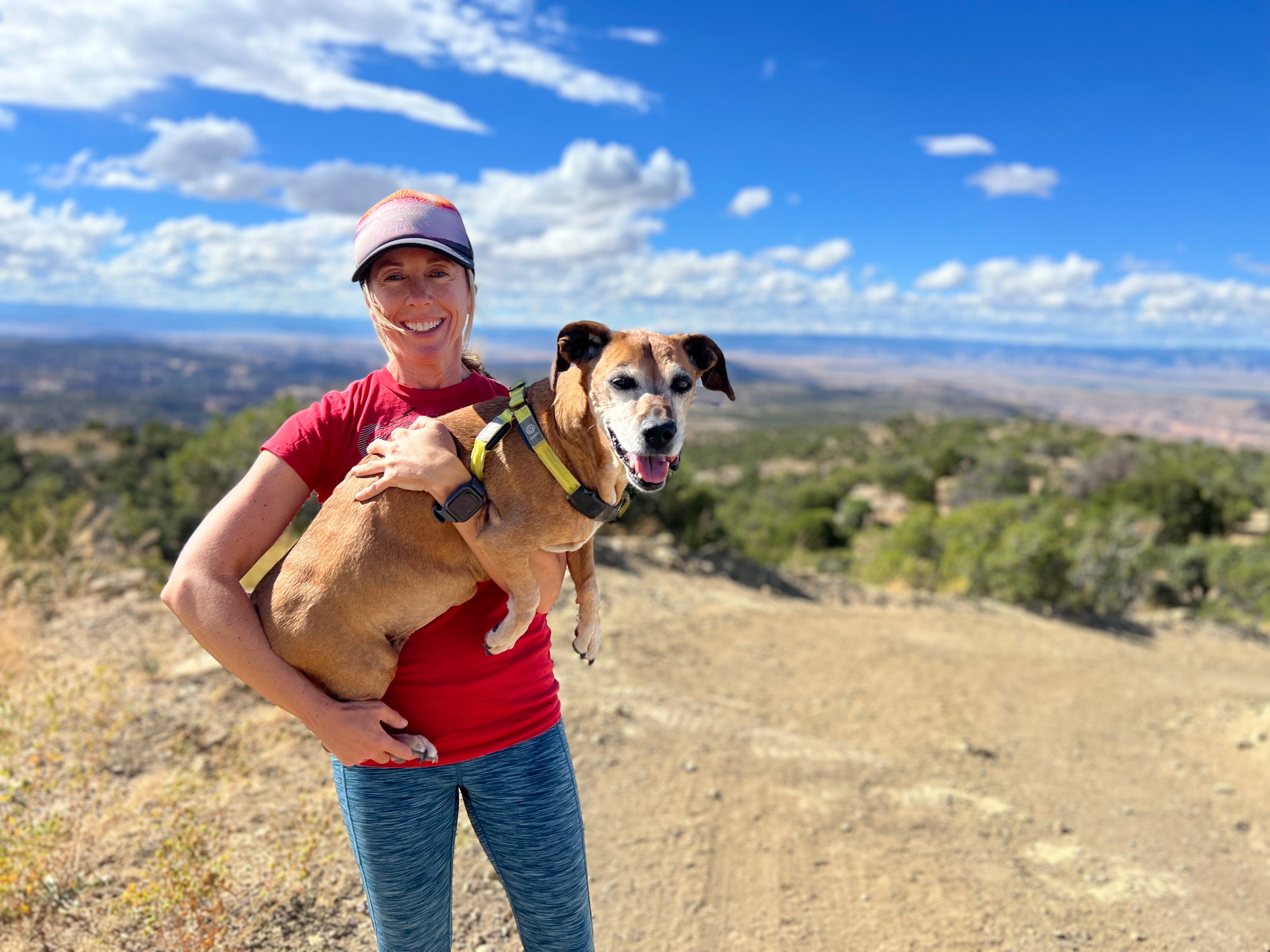 Smiling woman holding dog outdoors with scenic mountains, ideal for vanlife, road trip, adventure travel.