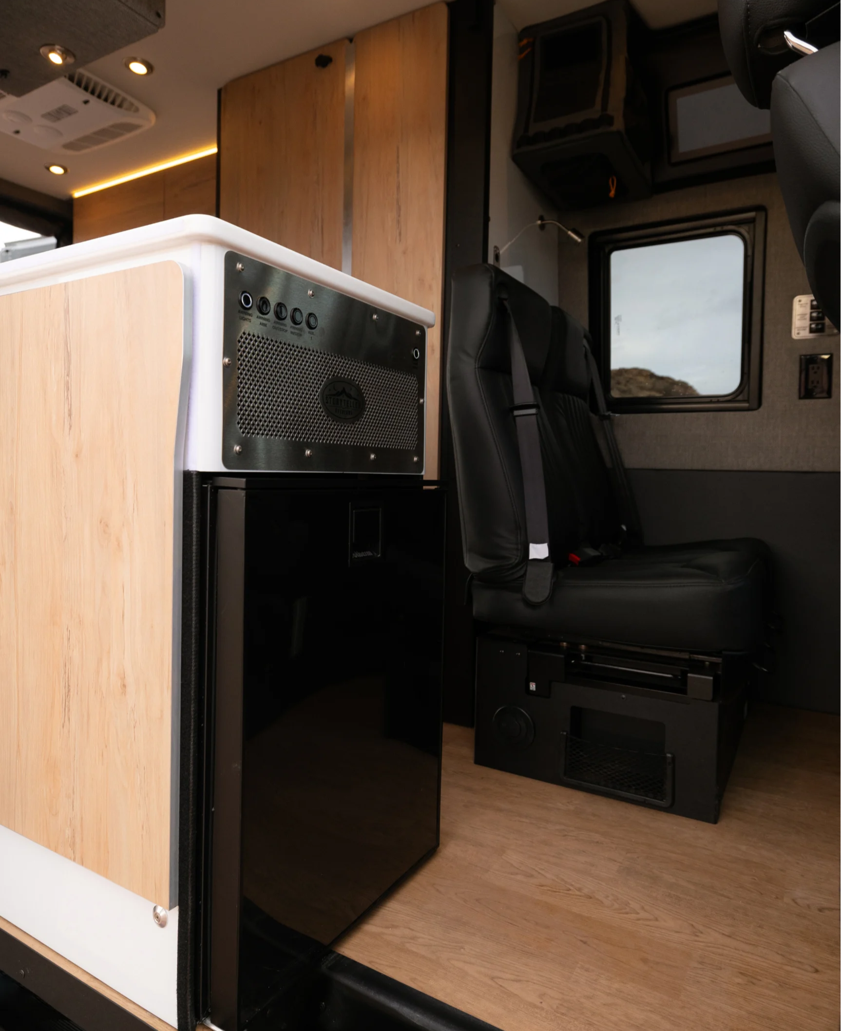 Modern camper van interior with wood panel cabinets, compact fridge, and black leather seats