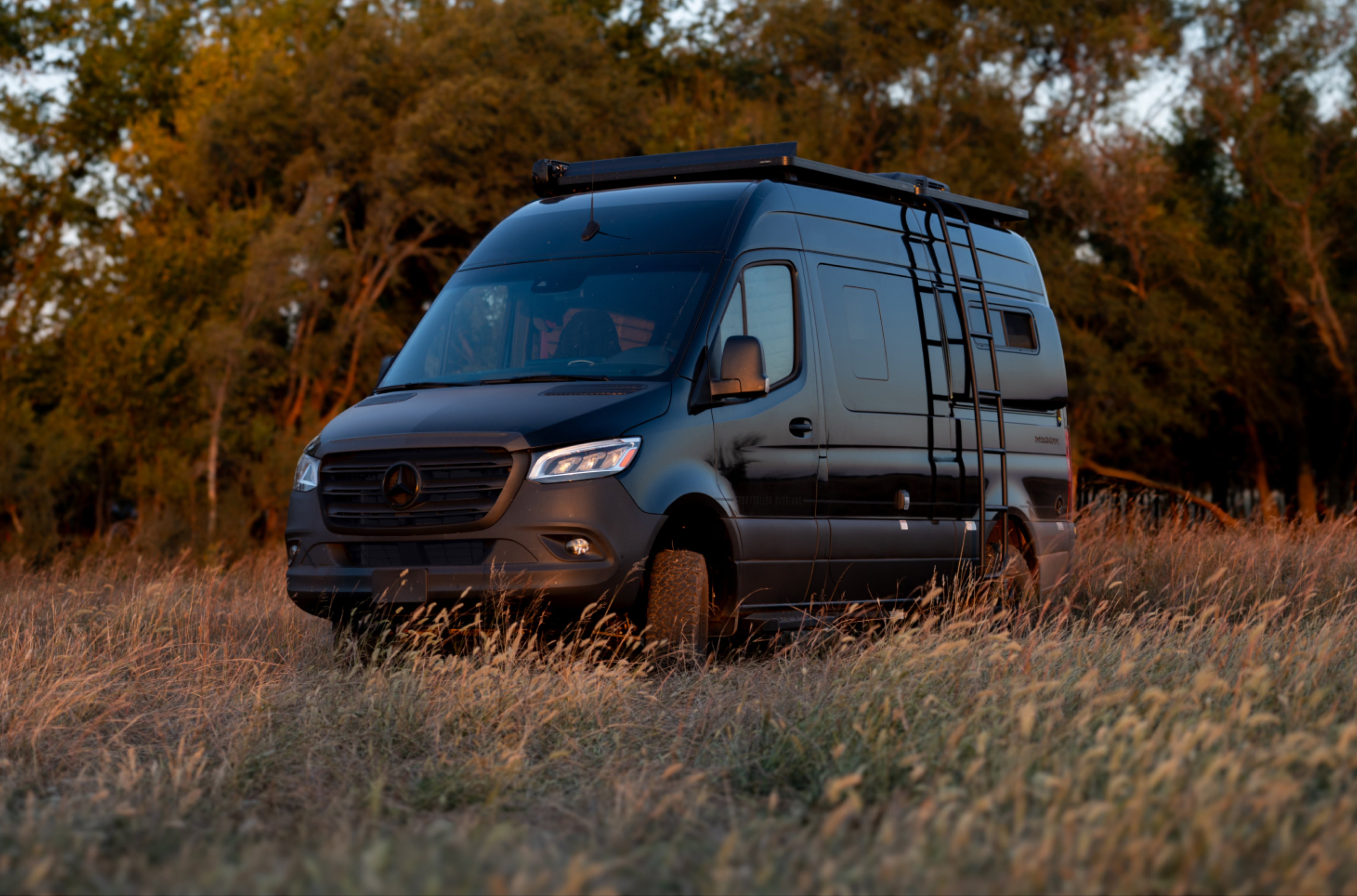 Stealth black Sprinter 4x4 camper van with roof rack and ladder in an offroad grassy field