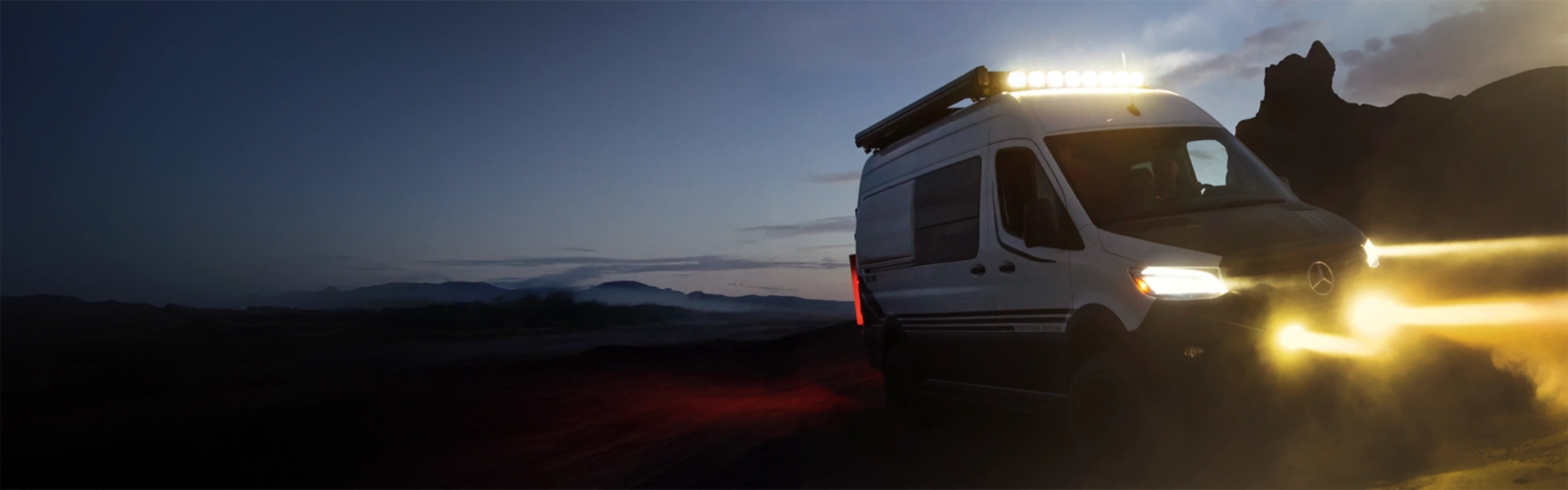 Adventure van Sprinter 4x4 campervan offroad at dusk with headlights on, mountain backdrop